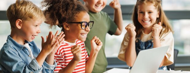 Schulkinder sitzen um einen Laptop und freuen sich über eine gemeisterte Aufgabe. 
